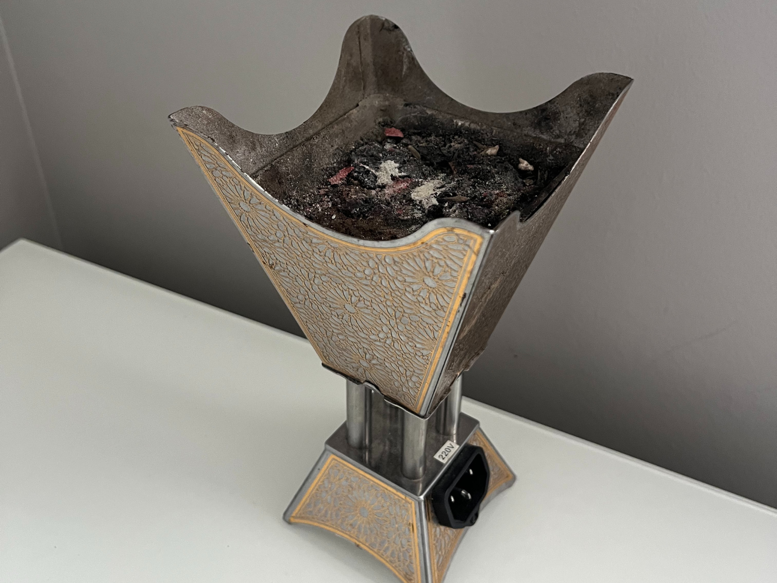 This is a photo of a traditional electric bukhoor burner, used for incense. It has a tall shape with flared edges at the top and a wide base for stability. The surface has golden floral designs that give it detail and cultural character. On the top, you can see ash and charcoal from incense that was burned. The photo is taken at a slight angle, so both the decorations and the inside depth are visible.