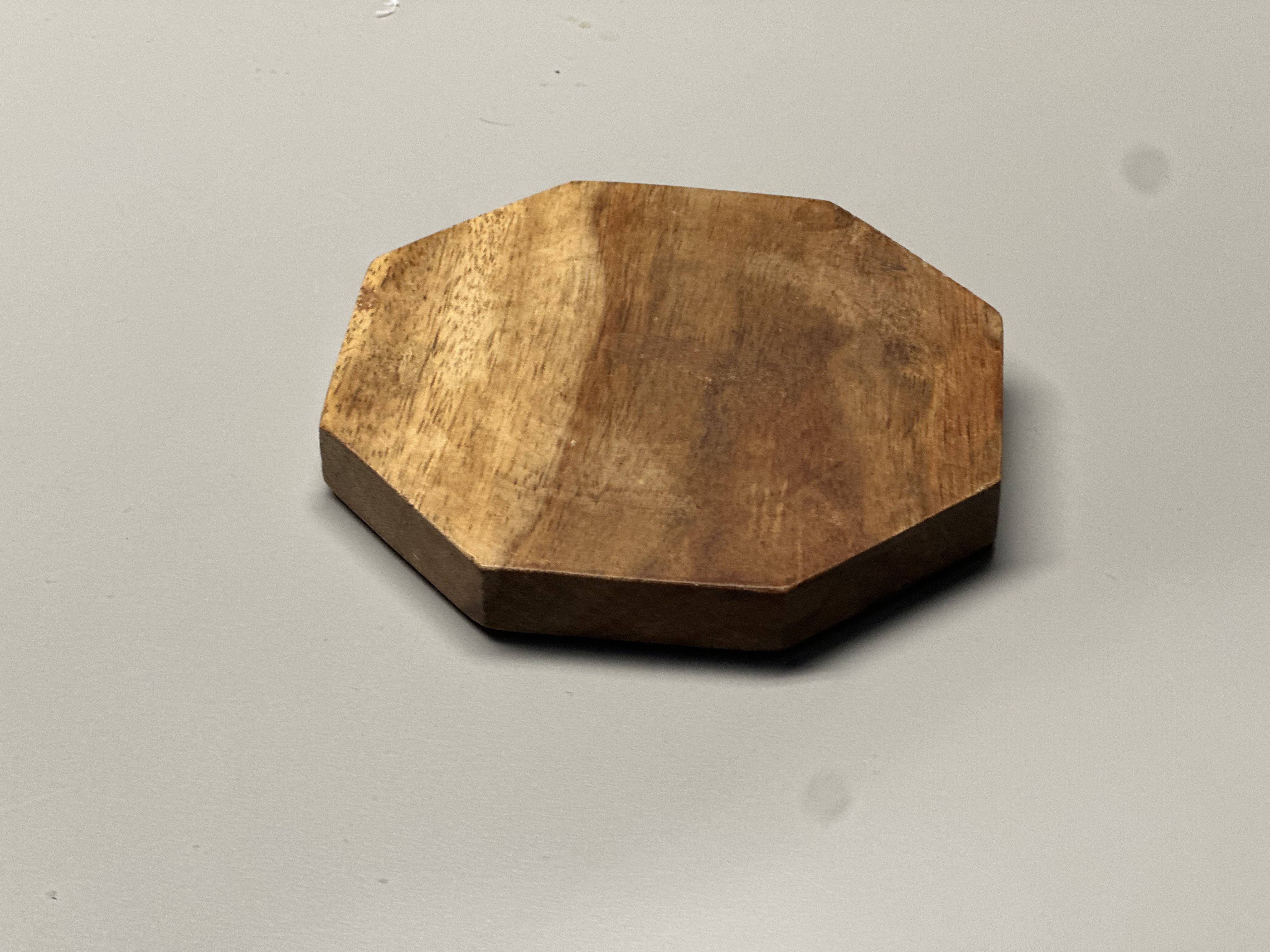 The material is wood that has been cut and sanded into an octagon shape. It is rigid, because wood is solid and holds its form. The surface looks semi-refined — it has been smoothed and shaped, but still shows natural grain patterns and slight texture. The warm brown color and visible wood fibers give it an organic and natural appearance.