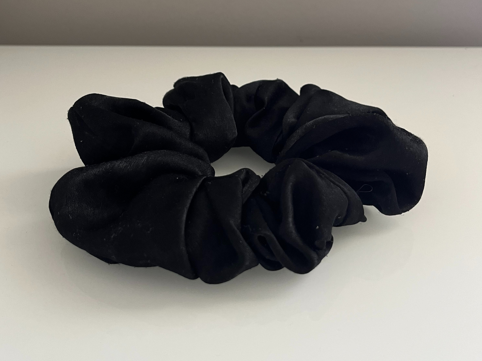This is another photo of the black scrunchie, taken from a lower side angle. This view shows more depth and volume in the folds, where the fabric bunches and overlaps. The angle also makes stronger shadows, which highlight the texture and shine of the material more than the top view.