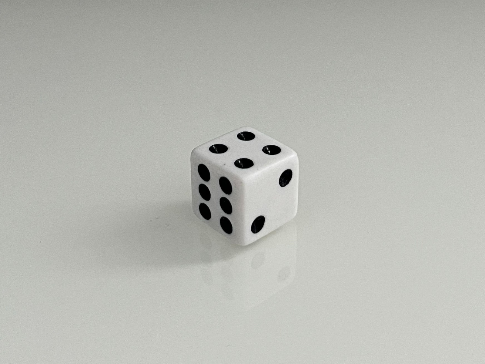 This is a photo of a white dice, taken at an angle where three faces are visible. The view shows the numbers four, five, and six, with the black dots standing out against the white surface. The light also makes a soft reflection under the dice, which gives the image more depth and realism.