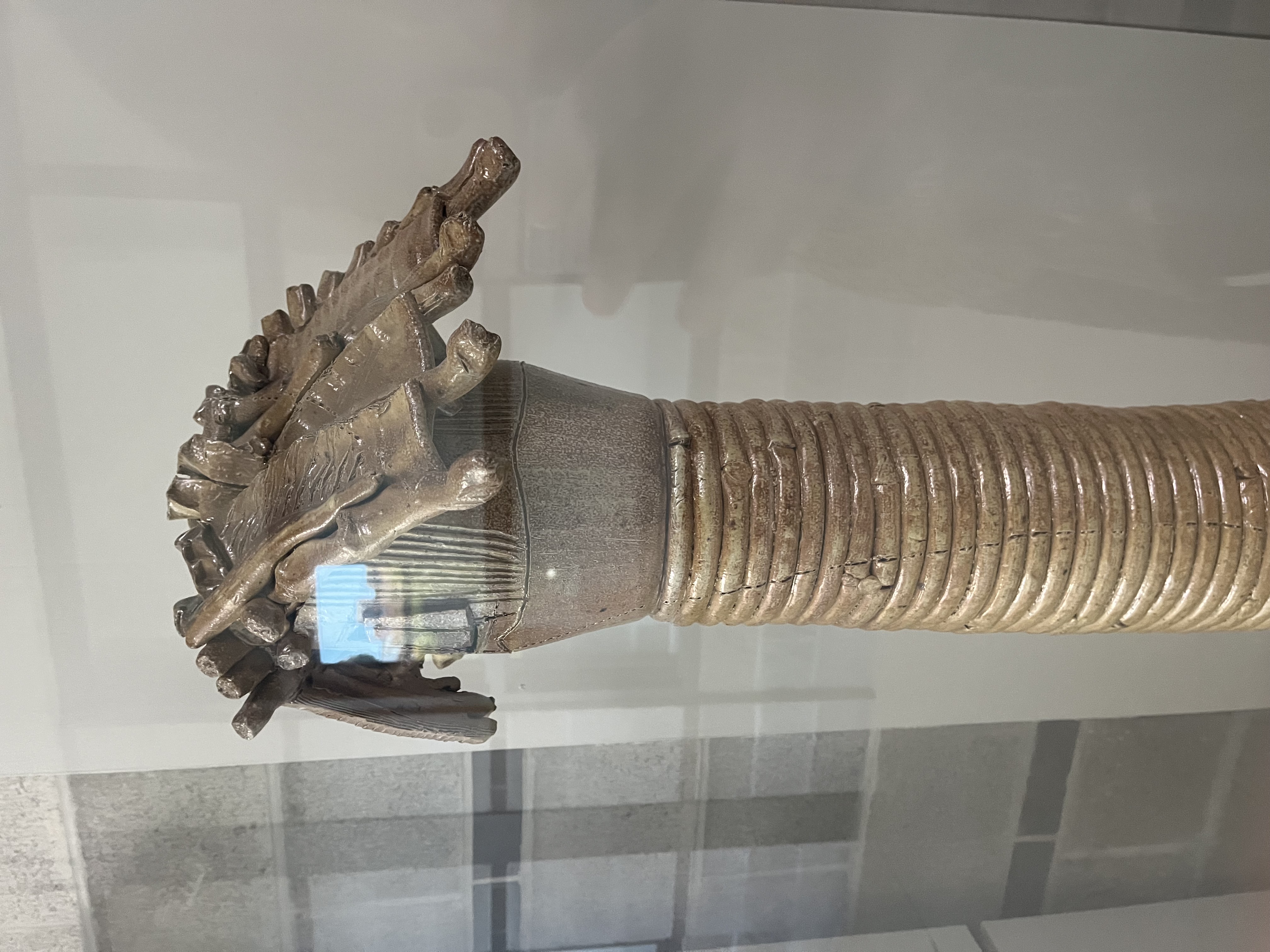 The sculpture is made from stoneware clay that has been glazed and fired. The material is rigid, because once clay is fired in a kiln, it becomes hard and solid. It is unrefined, with visible textures, cracks, and uneven surfaces that show the artist’s handwork. The glaze adds a slightly shiny and smooth finish on top, but the form still feels raw and natural. The piece looks handmade and expressive rather than perfect or polished.