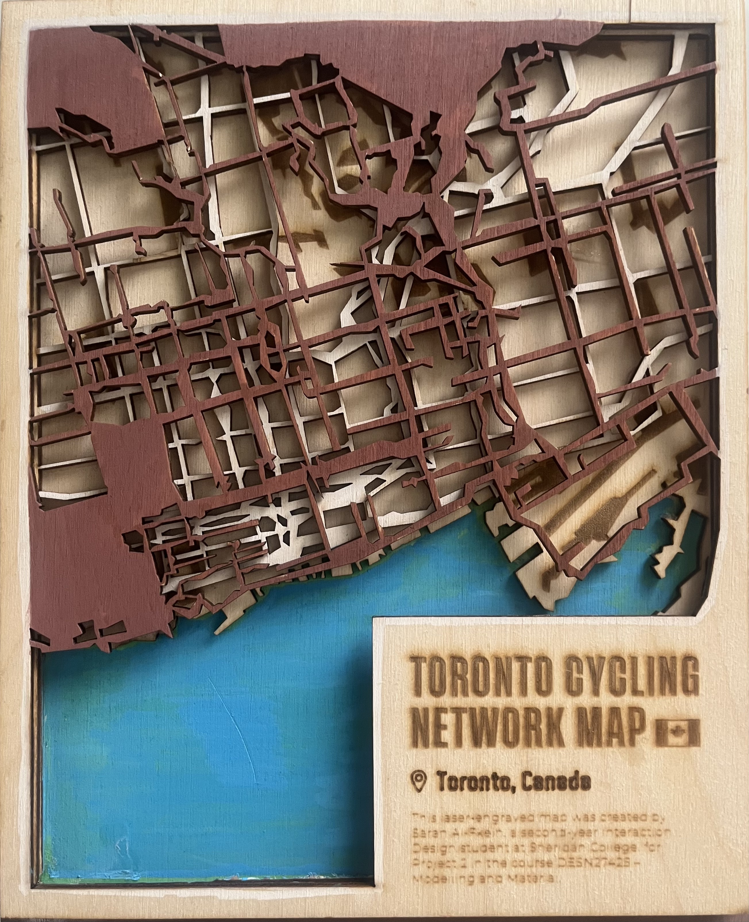 My final design is a multi-layered laser-engraved map of Toronto’s cycling network, created by exporting cycling-route data from Mapbox and translating it into vector layers suitable for fabrication. The piece is constructed from multiple sheets of birchwood, each representing a different type of spatial information: the base layer holds painted water features, the mid-layer shows Toronto’s landmass, and the top layer features the cycling routes engraved and cut with high precision. I hand-painted the water in blue and finished the cycling network in a warm brown tone to give the map a sense of depth, contrast, and clarity.

This project is deeply personal to me. I grew up in Saudi Arabia, where cycling—especially for women—was culturally discouraged and largely inaccessible. I never had the opportunity to learn as a child. When I moved to Canada at sixteen, I taught myself how to ride a bike through persistence and daily practice. After weeks of bruises, frustration, and determination, cycling became a new form of freedom for me. Today, it’s my favourite mode of transportation, especially in good weather.

Choosing Toronto as the focus felt natural. Toronto represents creativity, individuality, and a welcoming culture that contrasts strongly with where I grew up. It’s a city where I feel free to express who I am—through fashion, makeup, and my identity as a designer. I also regularly cycle in Toronto using public rental bikes, and I’ve always felt that the existing navigation apps don’t fully capture the logic of bike routes. By engraving the cycling network physically, I created a map that highlights the exact infrastructure cyclists rely on but is often hidden or simplified in digital maps.

The final engraved piece captures both the technical precision of laser fabrication and the emotional significance of cycling in my personal story. It represents my relationship with Toronto, movement, and belonging, while demonstrating the material craftsmanship and spatial thinking required in DESN 27425.